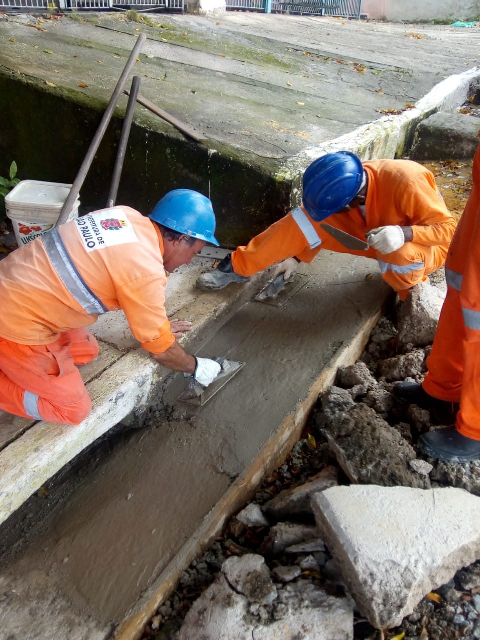 Dois trabalhadores uniformizados de laranja e capacete azul trabalham na reconstrução da sarjeta. Ao lado, pedaços de concreto que foram retirados. Os dois passam a desempenadeira, para deixar o concreto liso. 